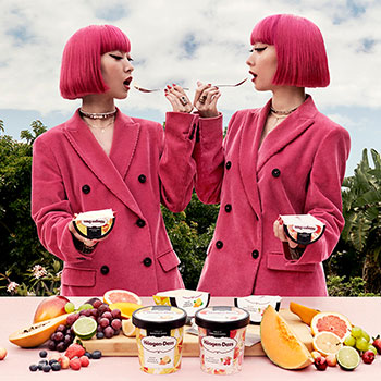 Two women with pink hair and coats eating in front of a table of fruit and two pints of Häagen Dazs ice cream
